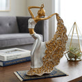 A decorative sculpture of a dancing woman with a flowing ornate skirt, shown on a wooden table in a styled living room setting with books and a plant.