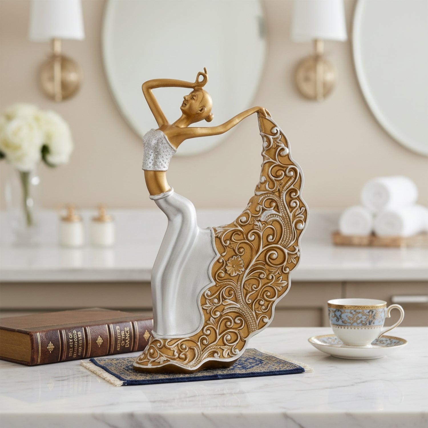 A decorative figurine of a dancing woman with a flowing ornate dress, shown on a tabletop in a softly styled interior setting with a book, teacup, mirrors, and candles in the background.