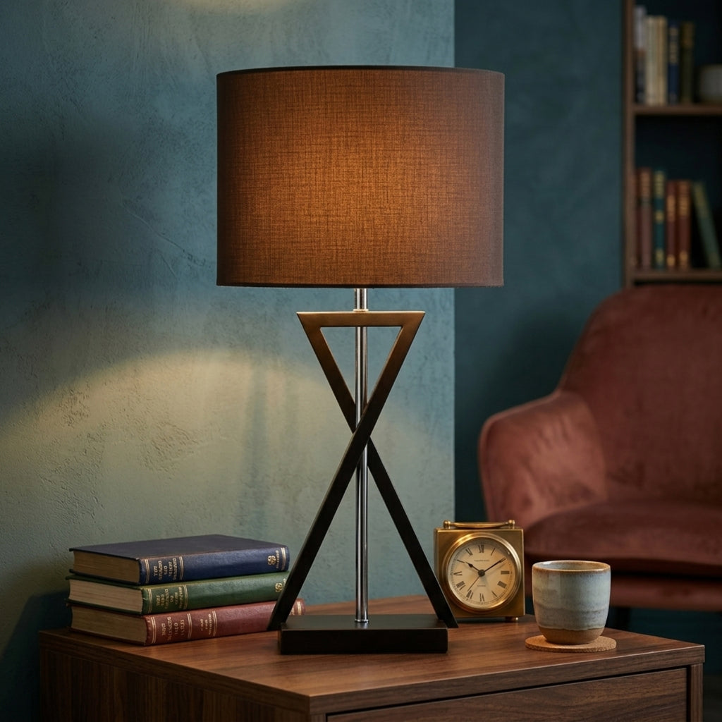 A table lamp with a brown fabric drum shade and a dark geometric metal base sits on a wooden side table in a styled room setting with books, a cup, and a chair.