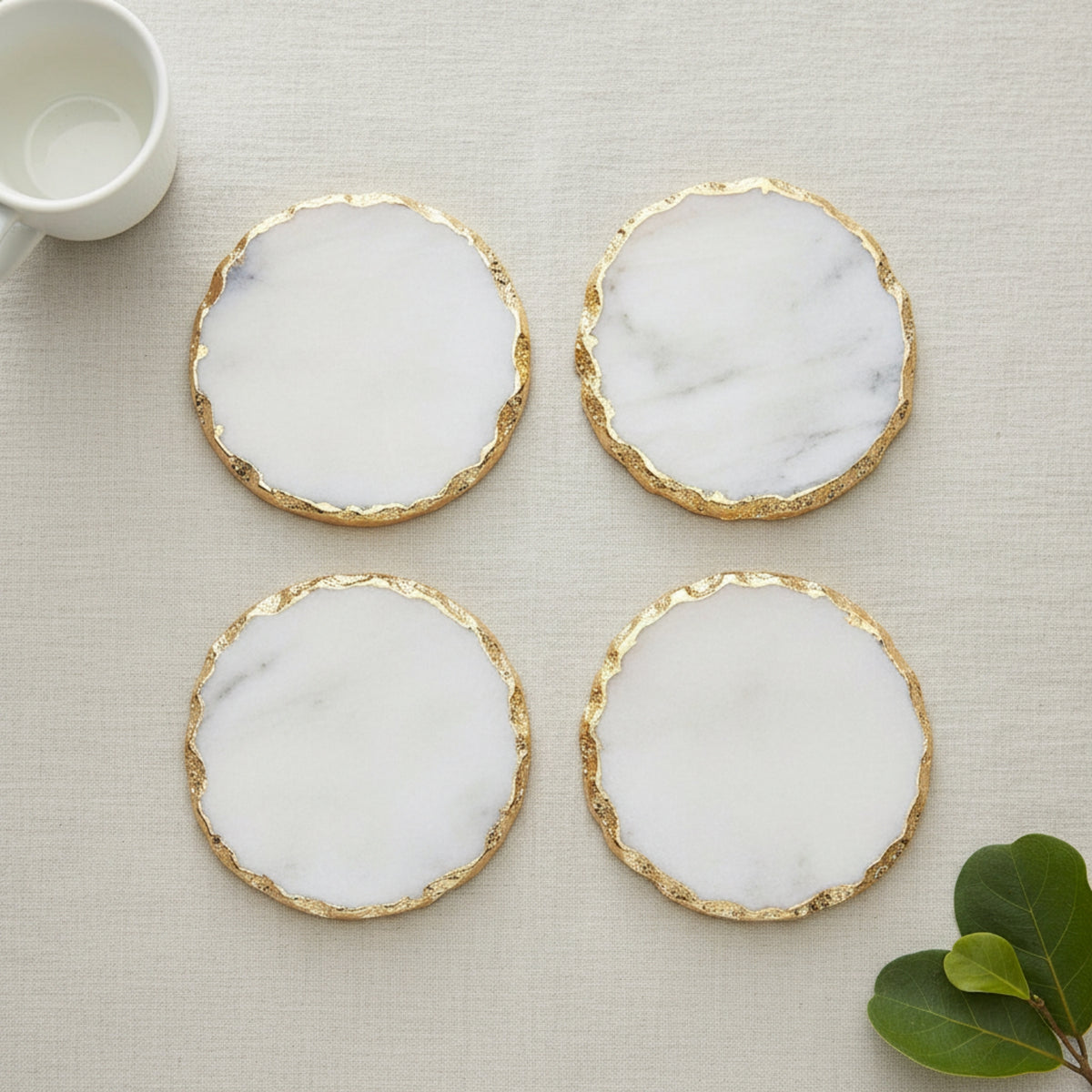 Four round coasters with white marble-look centers and textured gold edges, arranged in a 2x2 grid on a light beige fabric surface. A small white cup is partially visible in the top-left corner and green leaves appear in the bottom-right corner.