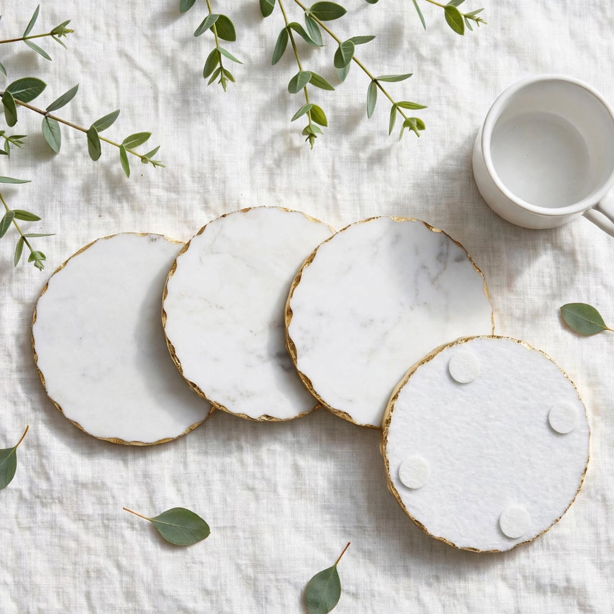 Four round white marble coasters with gold scalloped edges are arranged on a textured white linen surface, styled with scattered green eucalyptus leaves and a white ceramic mug in the upper right.
