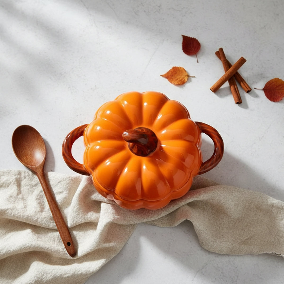 An orange pumpkin-shaped ceramic casserole dish with a lid and two handles, styled on a light stone surface with a wooden spoon, beige cloth, cinnamon sticks, and autumn leaves.