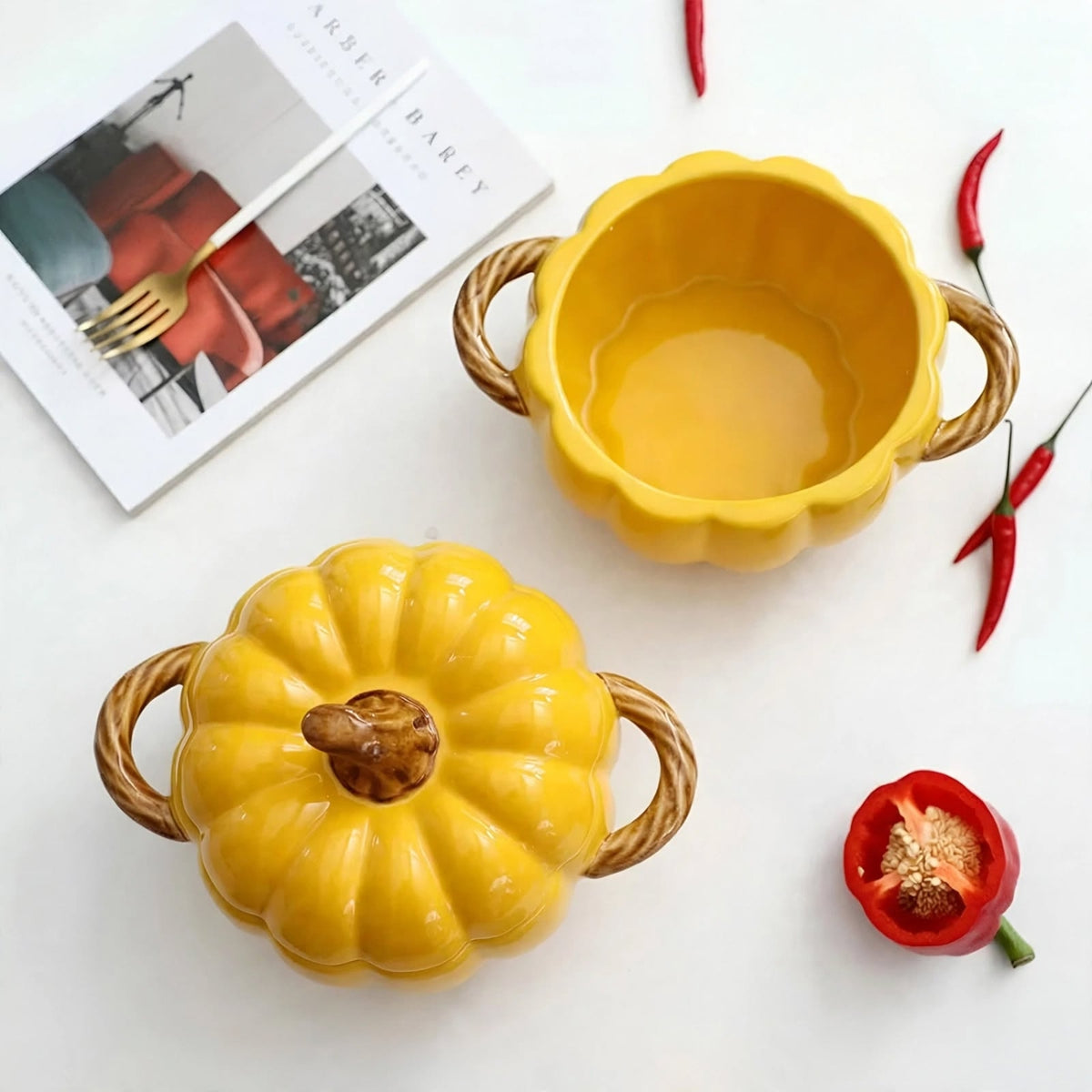 Two yellow ceramic pumpkin-shaped bowls with woven rope handles, one closed with a lid and one open, styled on a white surface with red chili peppers and a magazine.
