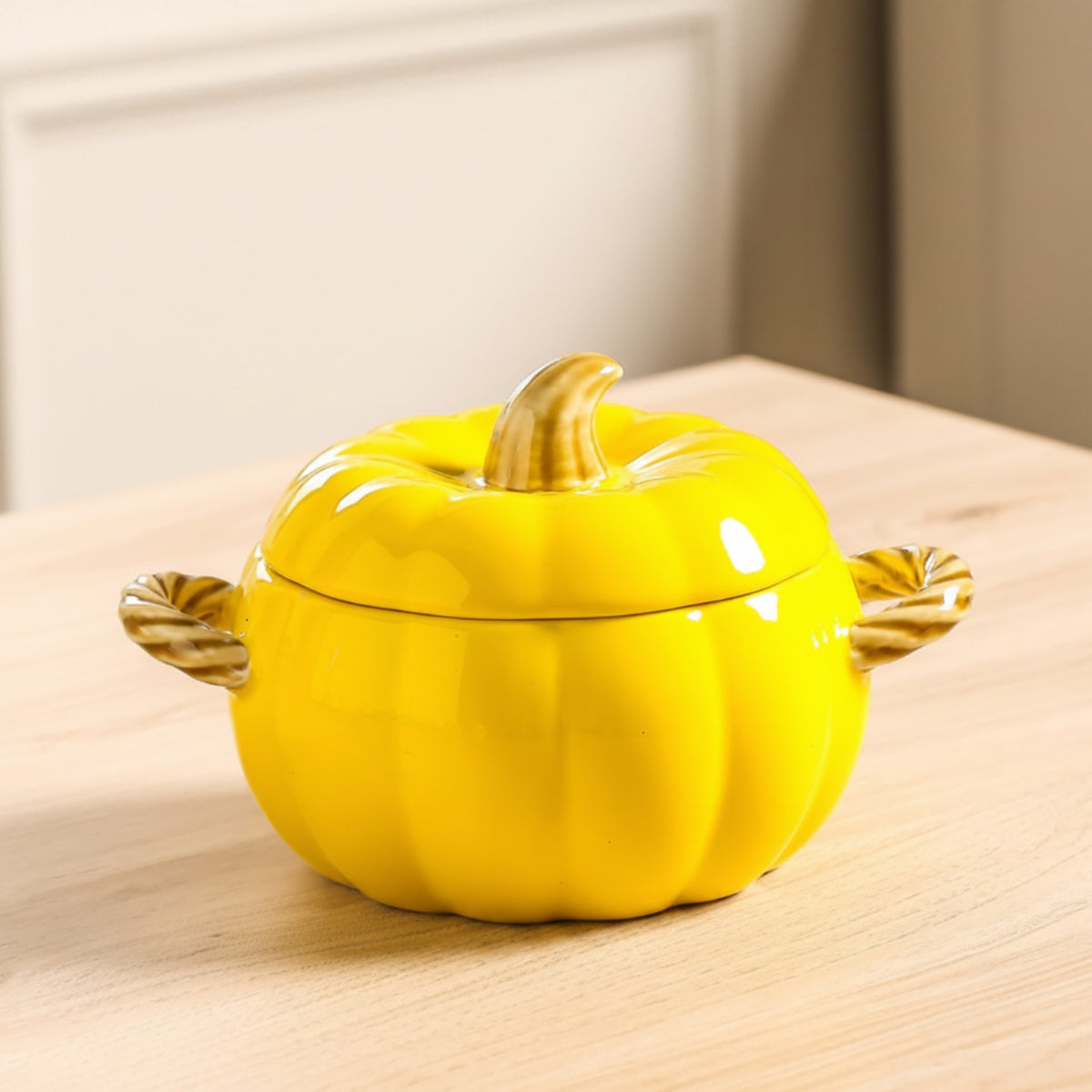 A bright yellow pumpkin-shaped ceramic lidded container with decorative twisted side handles and a faux stem, shown on a light wood tabletop in an indoor setting.