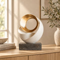 Modern abstract decorative sculpture with a white spiral shell and a metallic gold circular ring, displayed on a dark stone block base on a light wood console table in a styled interior setting.
