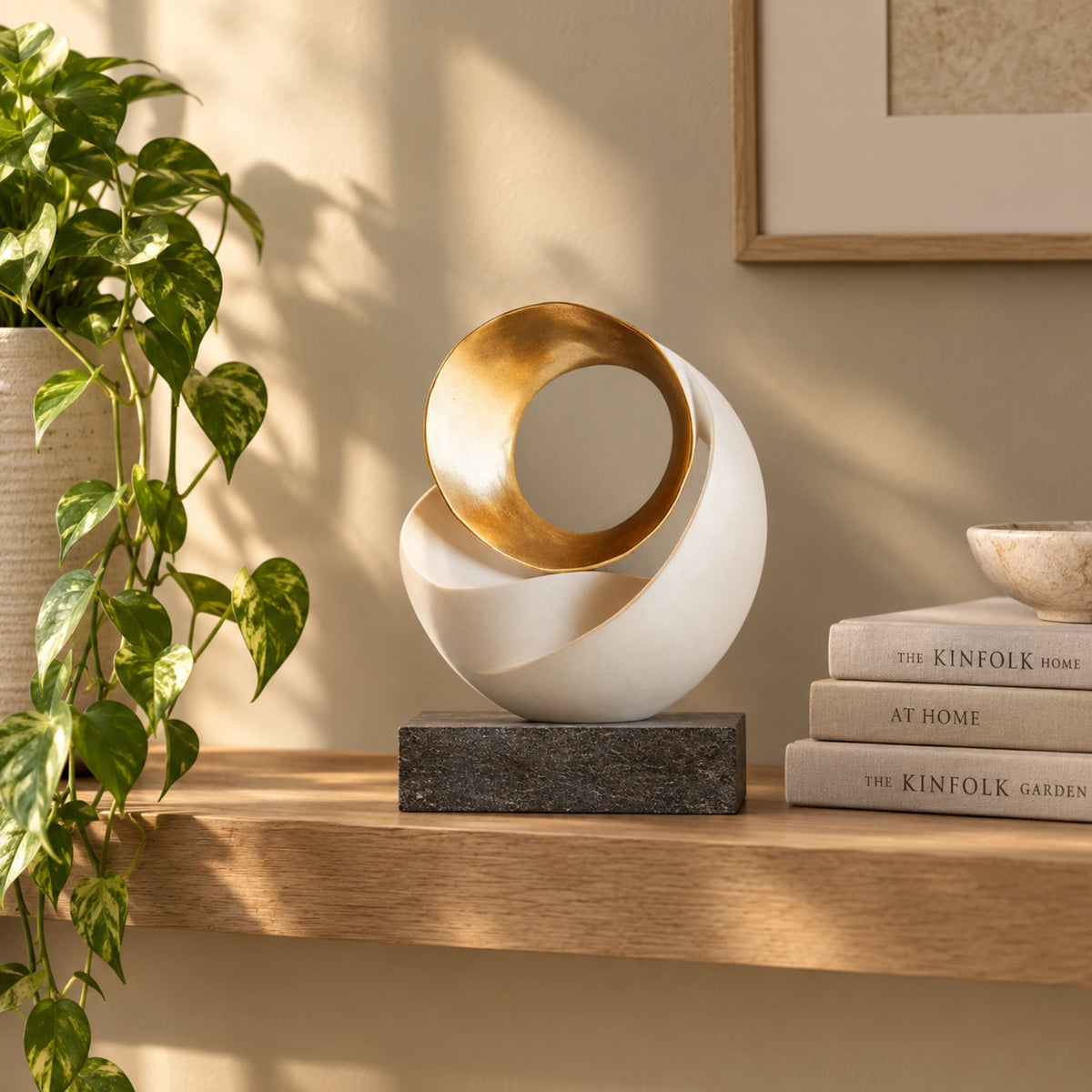 A modern abstract decorative sculpture with a white outer shell and a gold inner ring, displayed on a wooden shelf next to a green plant and stacked books in a warm, sunlit room.