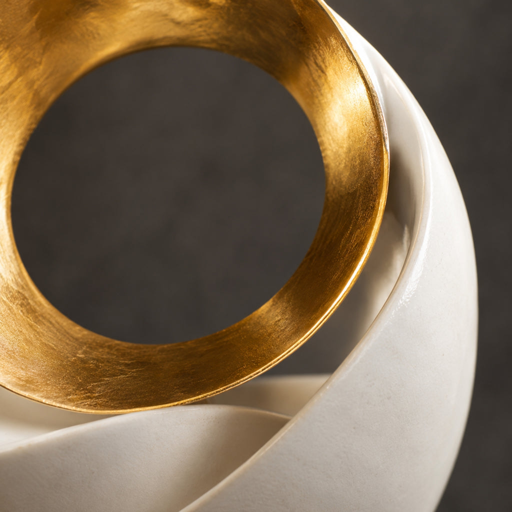 A close-up of a modern sculptural home decor piece featuring a matte white ceramic spiral shell with a textured, brushed gold metallic circular band resting on top, set against a dark gray background.