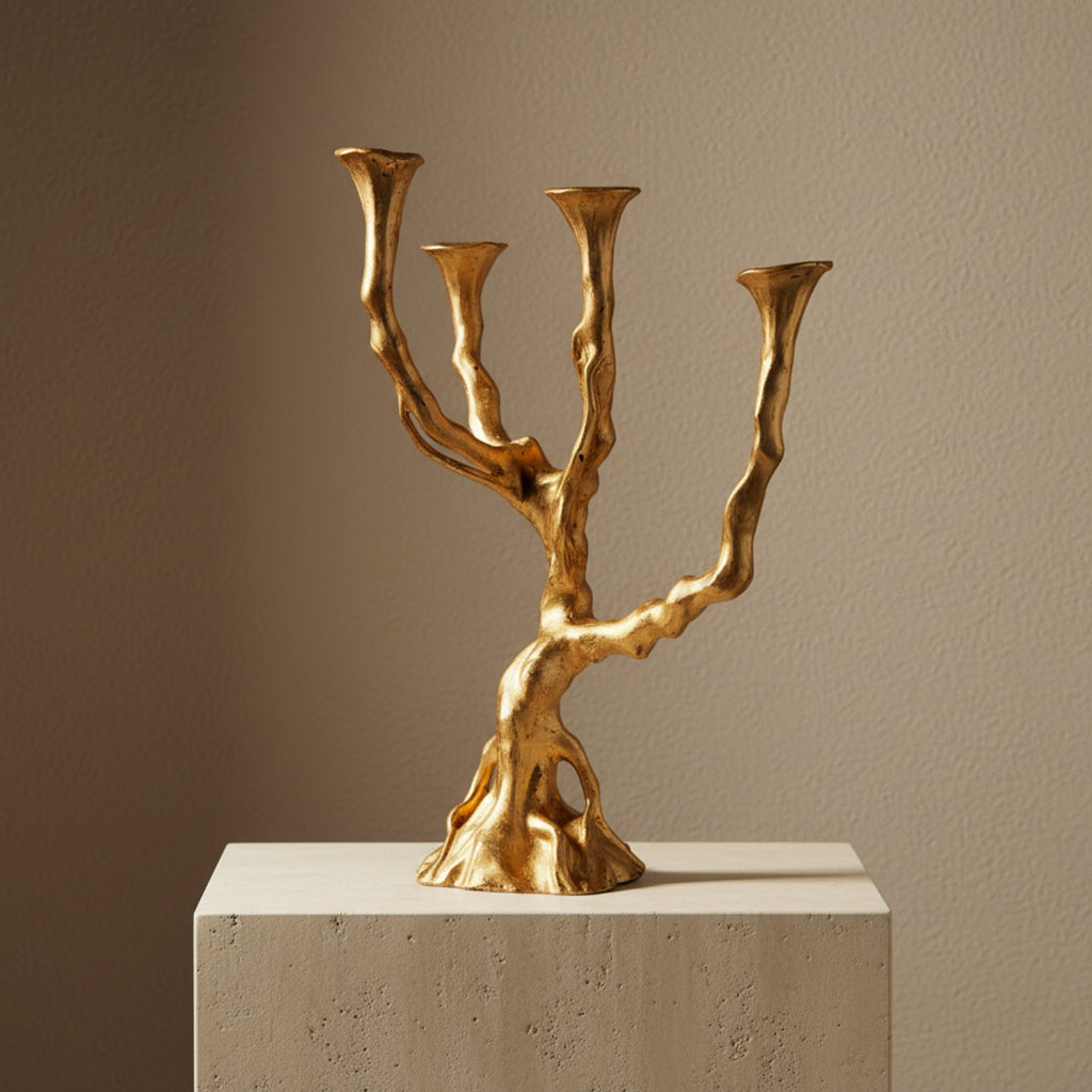 A three-arm gold candelabra styled like a gnarled tree trunk, displayed on a textured white pedestal against a warm beige wall.