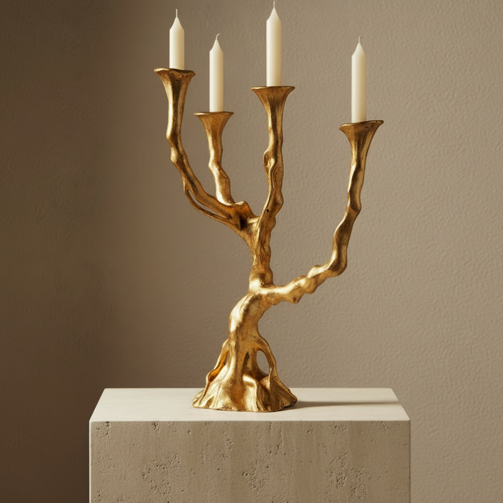 A sculptural gold candelabra with four candle holders shaped like gnarled tree branches, shown with four white candles on top of a light stone pedestal against a warm taupe wall.