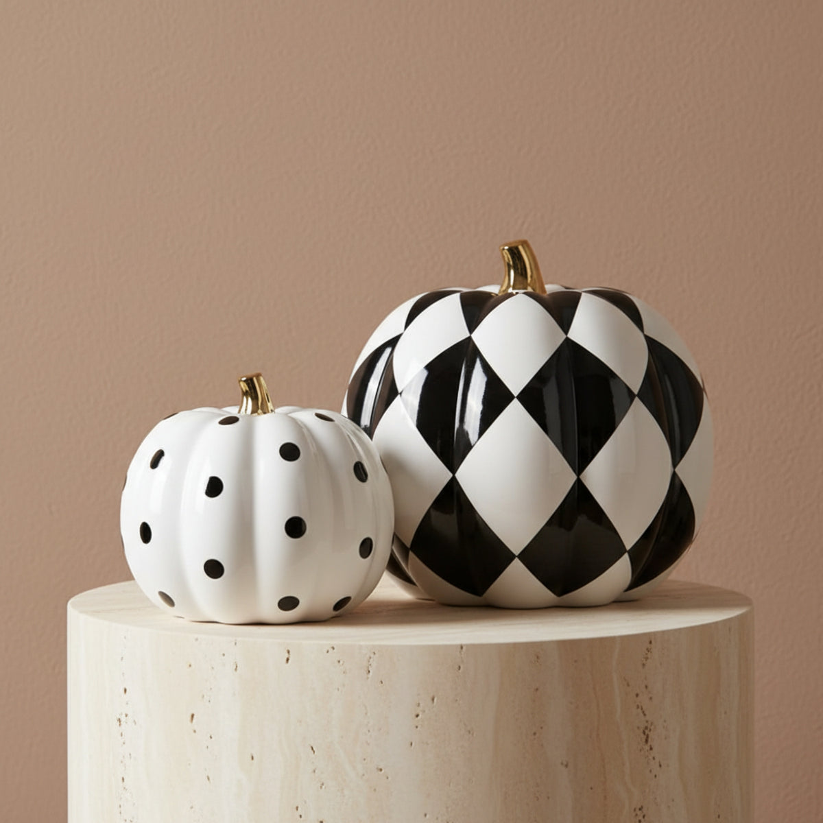 Two decorative ceramic pumpkins displayed on a round pedestal against a warm beige wall. The larger pumpkin has a black and white harlequin diamond pattern with a small gold stem, and the smaller pumpkin is white with black polka dots and a gold stem.