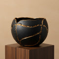 A black ceramic bowl with a cracked glaze design highlighted by metallic gold lines, displayed on a dark wood pedestal against a warm beige background.