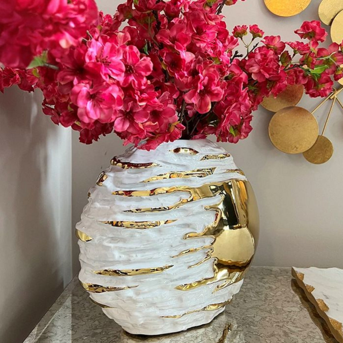 A white ceramic vase with textured horizontal ridges and metallic gold accents, filled with bright pink artificial flowers, displayed on a marble-like surface against a neutral wall.