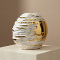 A sculptural oval vase with a textured white ceramic body and reflective gold metallic accents, displayed on a light stone pedestal against a warm beige wall.