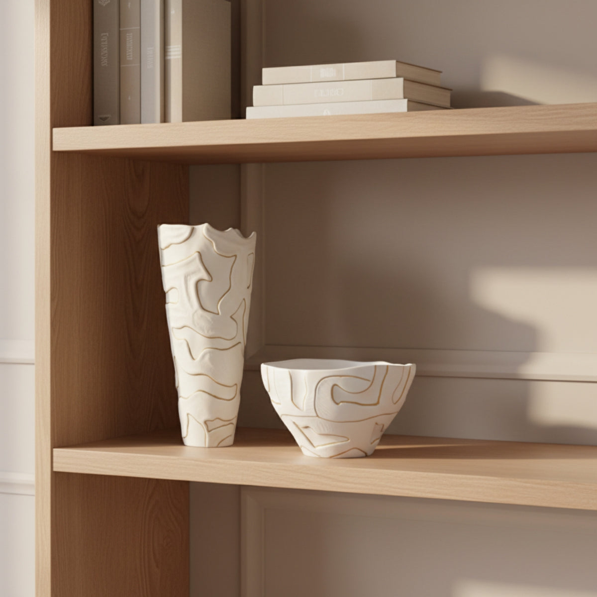 A white ceramic vase and matching bowl with abstract gold line patterns, displayed on a light wood bookshelf next to books.