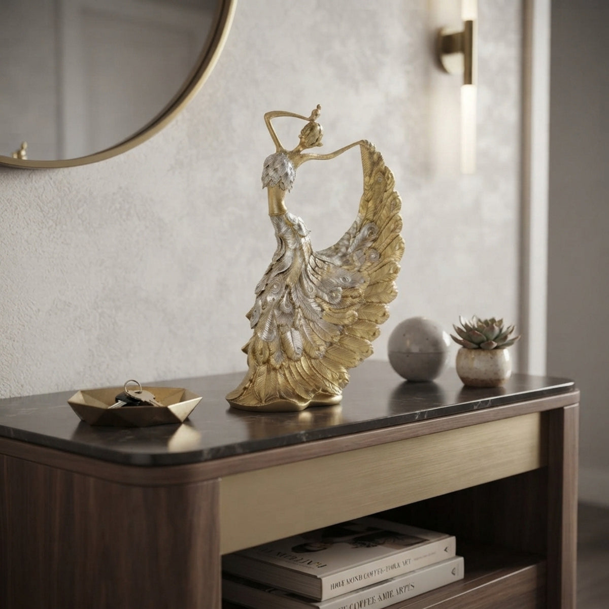 A decorative gold and silver dancer figurine stands on a dark marble-topped console table in a styled interior setting with a mirror, wall sconce, small tray, and succulent.