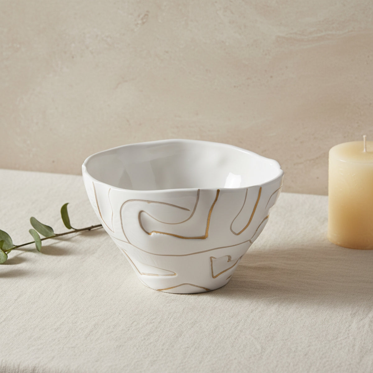 A white ceramic bowl with abstract gold line patterns, styled on a textured beige surface with a candle and eucalyptus sprig in the background.