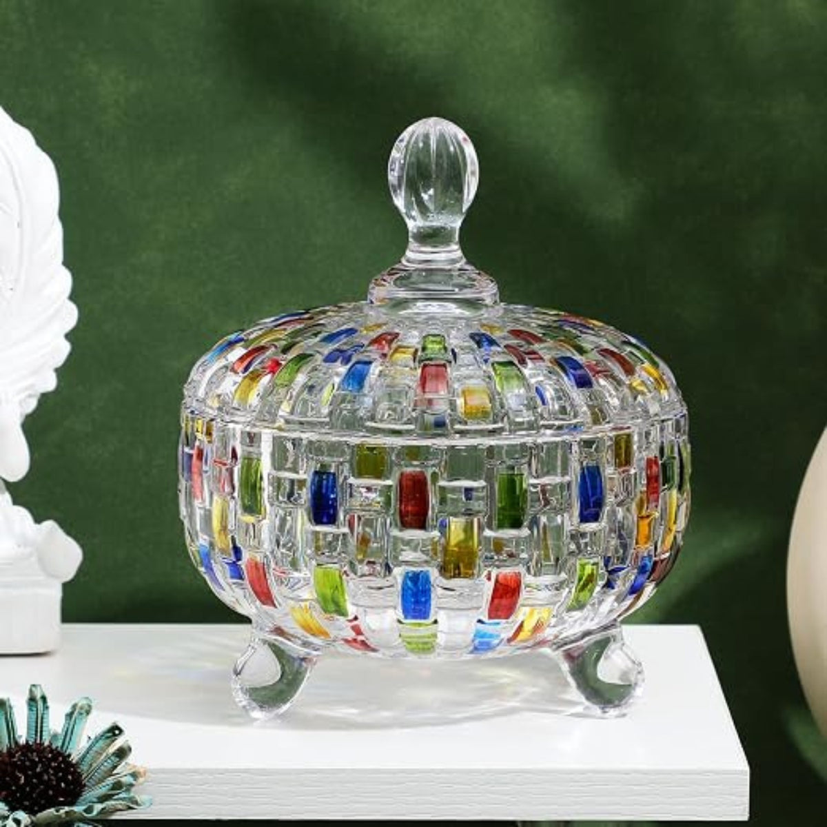 A clear glass lidded candy dish with colorful mosaic tile accents on the body and lid, shown on a white surface against a dark green backdrop.