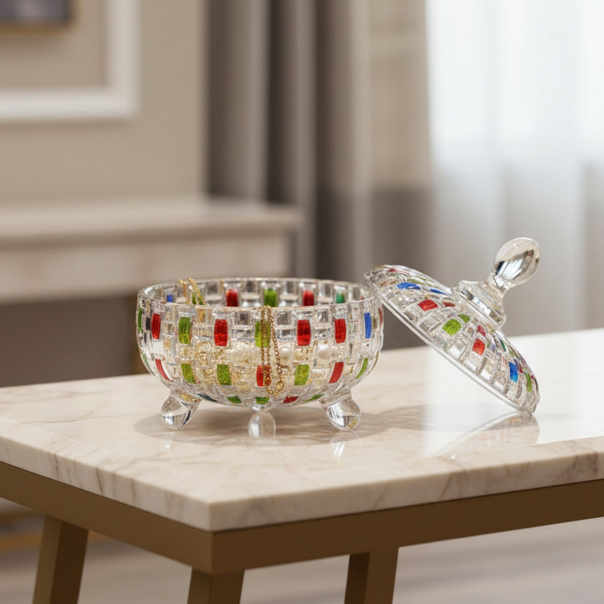A clear glass decorative bowl with a matching lid, featuring colorful mosaic accents in red, green, and blue, displayed on a marble-topped table in a softly lit indoor setting.