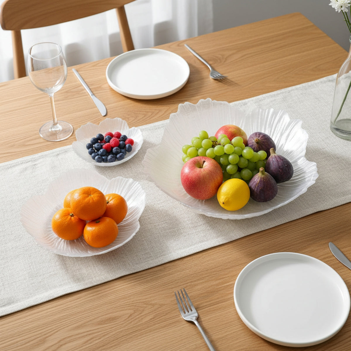 A styled dining table setting featuring three white scalloped ceramic fruit bowls arranged on a beige table runner. The bowls contain oranges, blueberries, and a mixed fruit assortment including grapes, apples, and figs, set against a light wood table with white plates and silver cutlery.