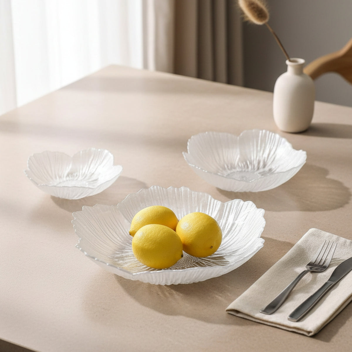 Three clear ribbed glass bowl plates arranged on a beige table, with one bowl in the foreground holding two yellow lemons. A folded cream napkin with silver fork and knife sits at the lower right, and a small white vase with dried stems is in the background near a window.