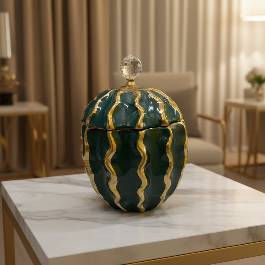A decorative lidded storage jar with a rounded, ribbed design in glossy dark green and gold, topped with a clear crystal knob, displayed on a white marble table in a modern living room setting.