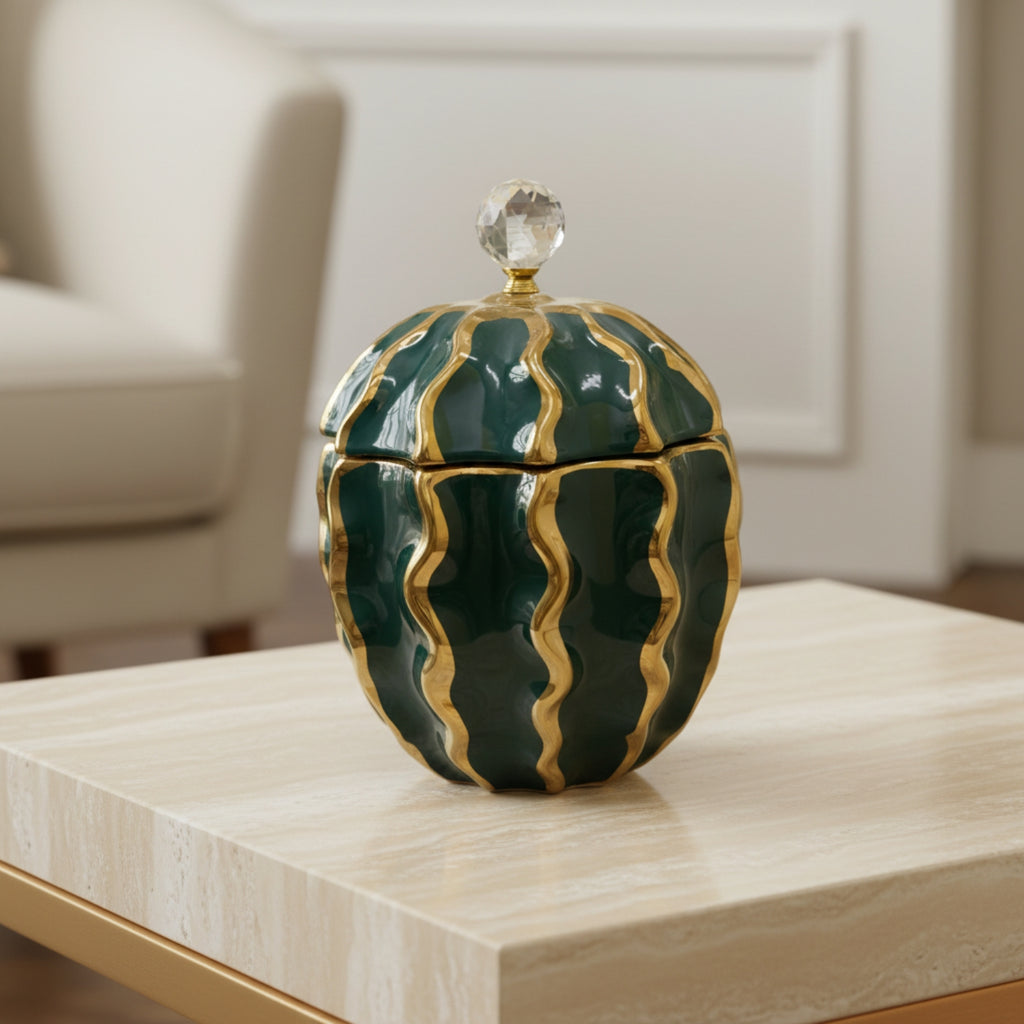 A decorative lidded container with a rounded, ribbed design in glossy dark green and gold, topped with a clear crystal knob, displayed on a light wood table in a modern living room setting.