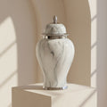 A white and gray marble urn with a silver metal lid and base, displayed on a beige pedestal in a softly lit interior setting.