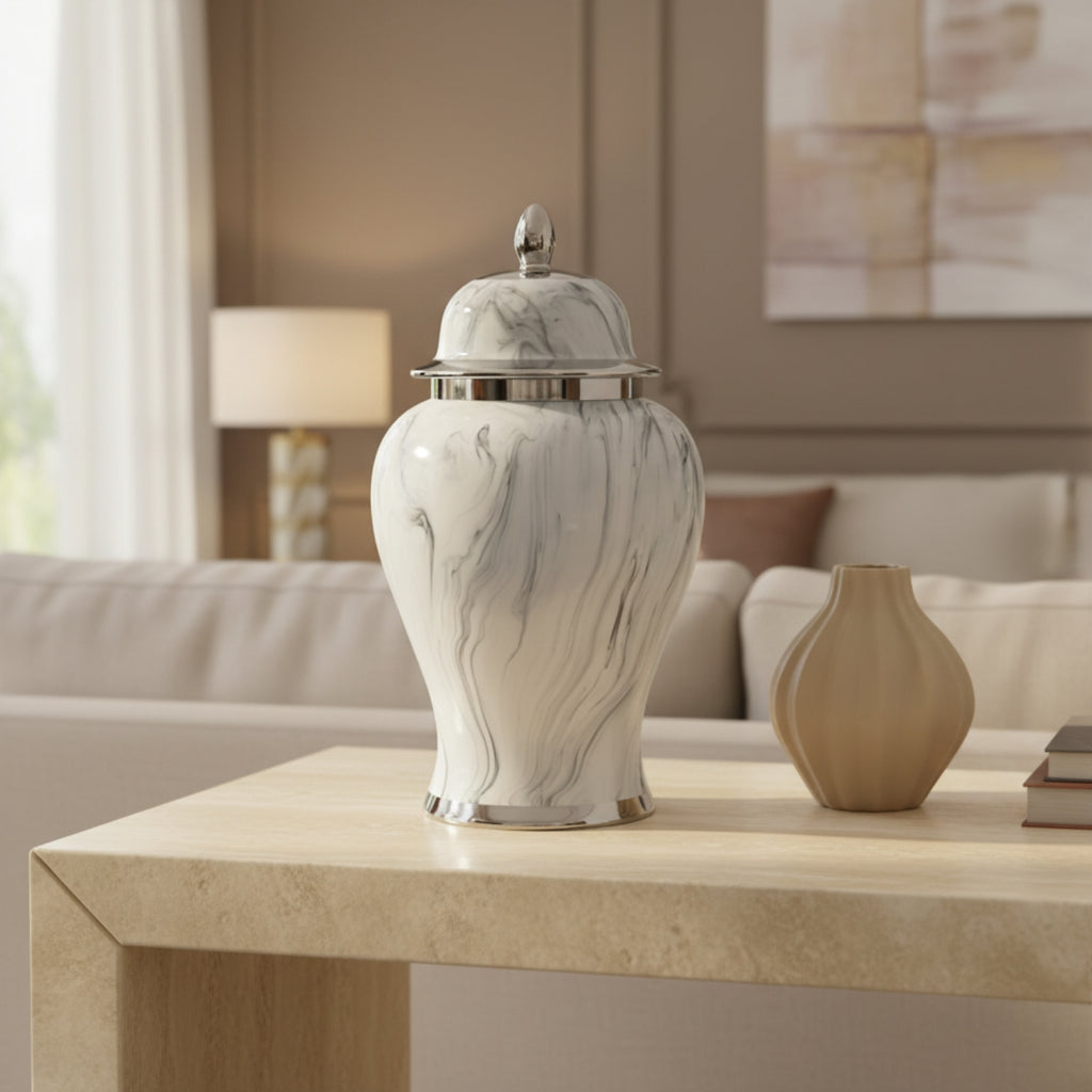 A white and gray marble-patterned lidded urn with silver trim sits on a light wood table in a modern living room setting, next to a small beige vase.