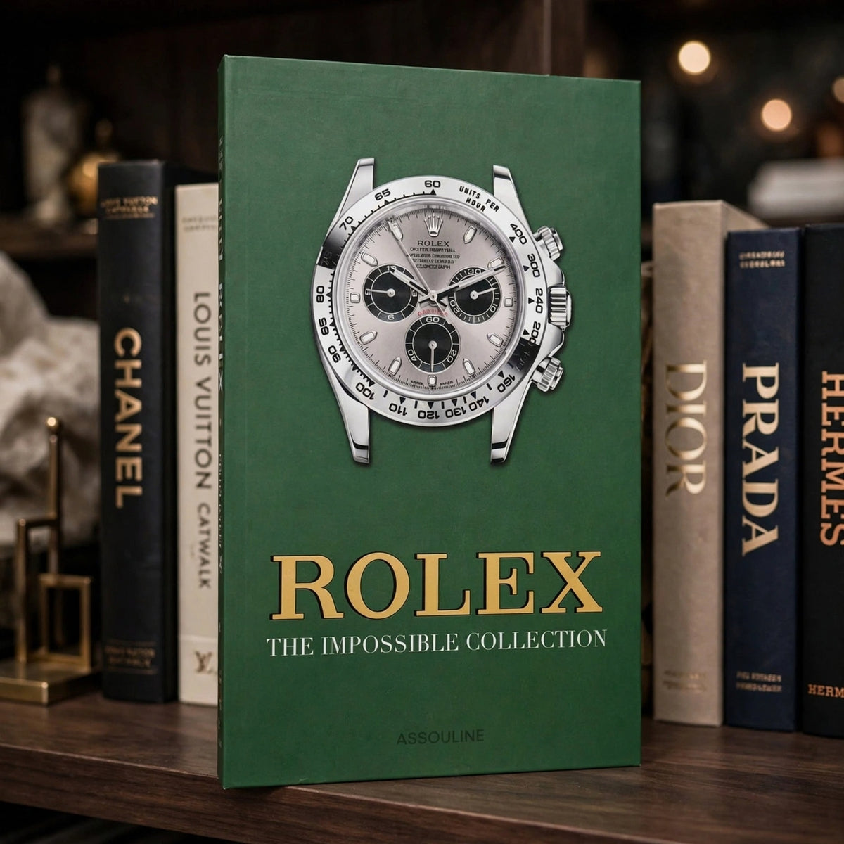 A green hardcover book titled 'Rolex: The Impossible Collection' by Assouline, featuring a large Rolex watch face on the cover, standing on a wooden shelf between other luxury books including Louis Vuitton, Chanel, Dior, Prada, and Hermès.