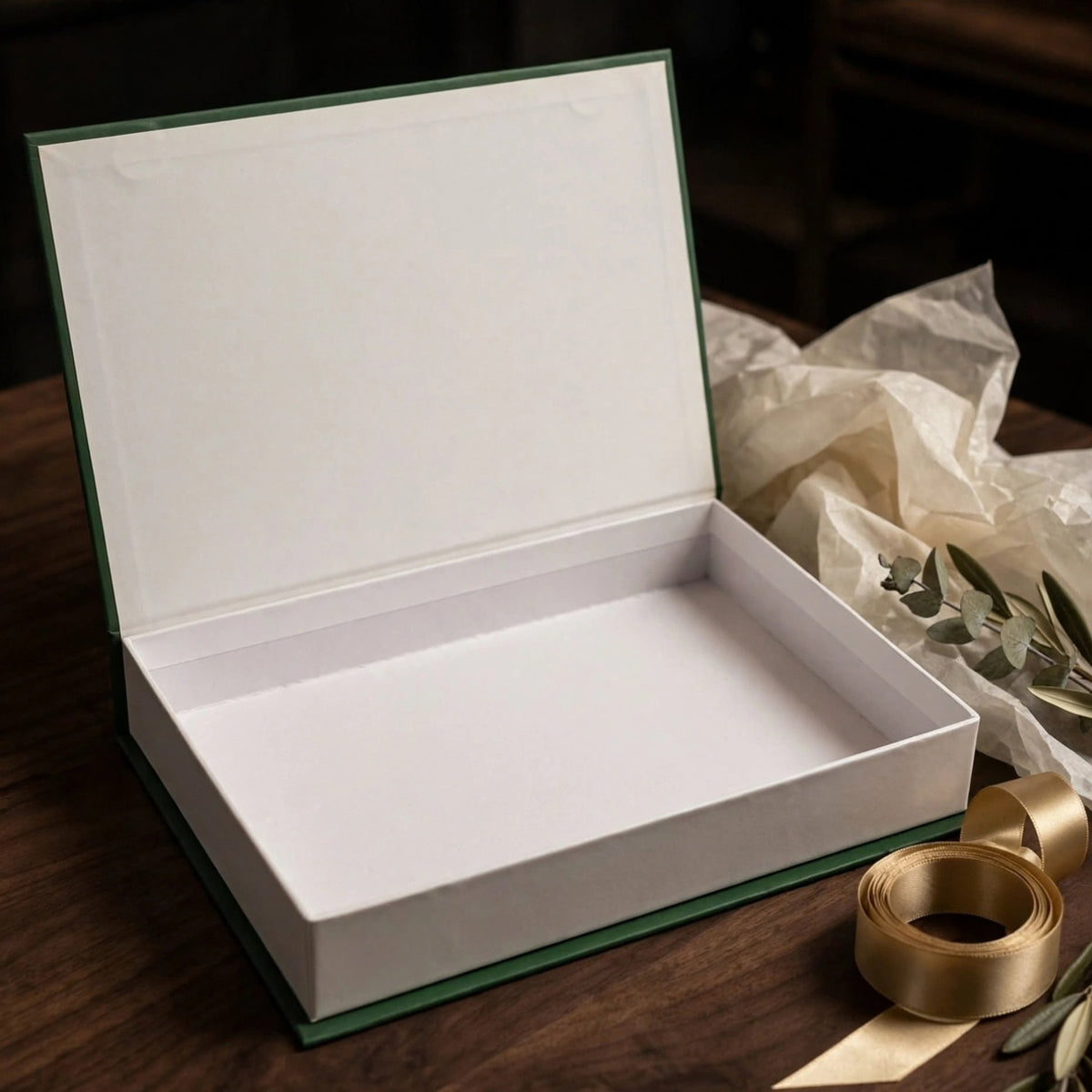 An open rectangular gift box with a white interior and dark green edges, placed on a wooden surface with crumpled tissue paper and a gold ribbon nearby.