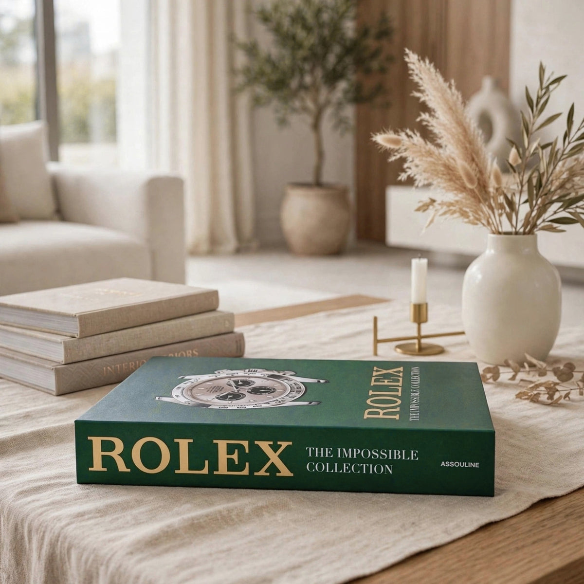 A green Rolex hardcover book titled 'The Impossible Collection' by Assouline, styled on a beige linen tablecloth in a bright, modern living room setting.