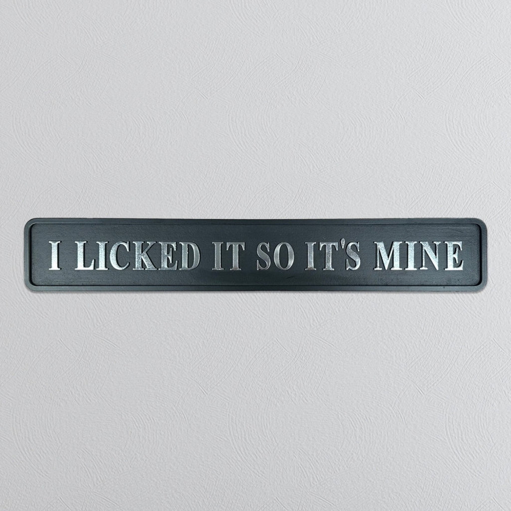 A rectangular dark gray sign with raised silver lettering that reads 'I LICKED IT SO IT'S MINE' on a light gray textured background.