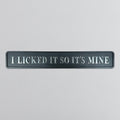 A rectangular dark gray sign with raised silver lettering that reads 'I LICKED IT SO IT'S MINE' on a light gray textured background.