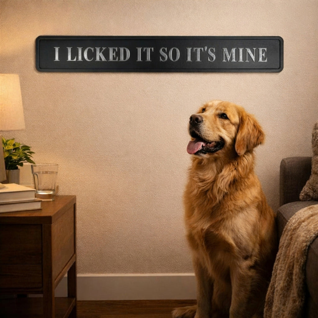 A golden retriever dog sits in a cozy living room next to a sofa, looking up at a black rectangular wall sign that reads 'I LICKED IT SO IT'S MINE' in white lettering.