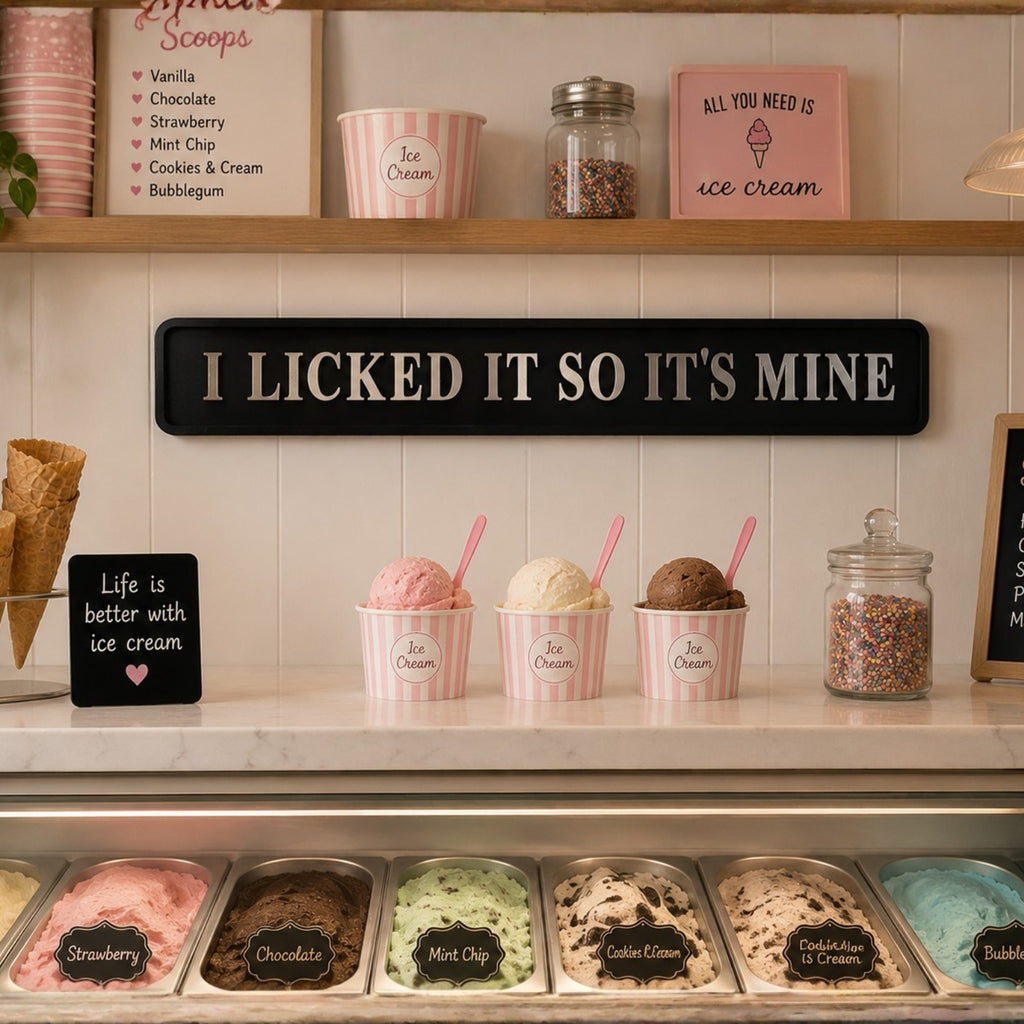 A decorative black wooden sign with white text reading 'I LICKED IT SO IT'S MINE' mounted on a cream-colored wall above an ice cream display. The scene includes pink-striped ice cream cups with pink spoons, jars of sprinkles, and multiple flavors in metal trays below.