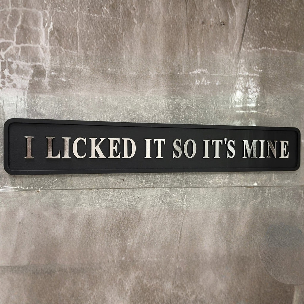 A rectangular black sign with raised silver lettering that reads 'I LICKED IT SO IT'S MINE' mounted against a textured gray stone wall.