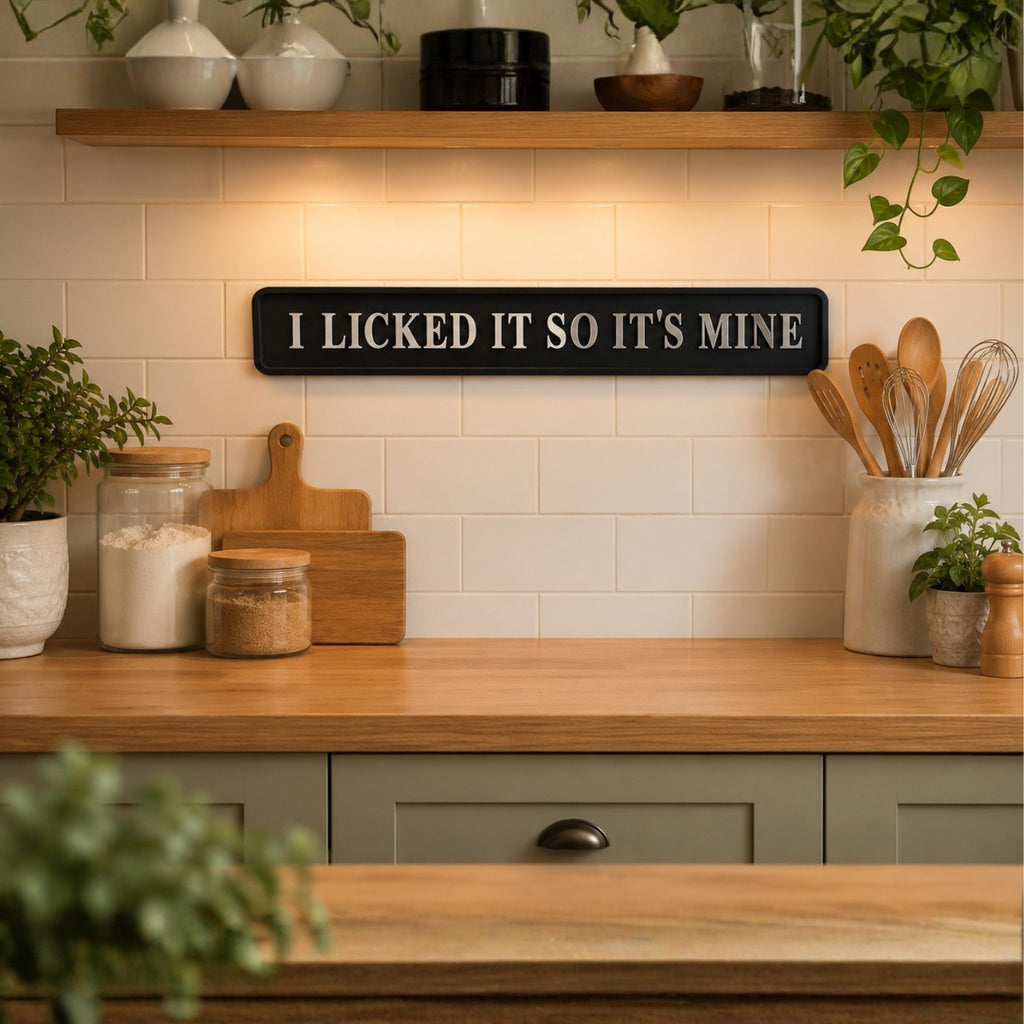 A black rectangular wall sign with white text reading 'I LICKED IT SO IT'S MINE' mounted on a cream subway tile kitchen backsplash above a wooden countertop with jars, cutting boards, and utensils.