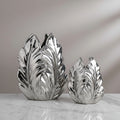 Two metallic silver leaf-shaped decorative vases of different sizes, displayed on a white marble surface against a neutral gray background.