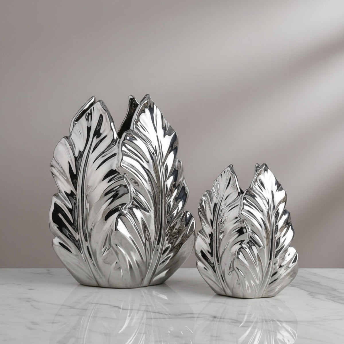 Two metallic silver leaf-shaped decorative vases of different sizes, displayed on a white marble surface against a neutral gray background.