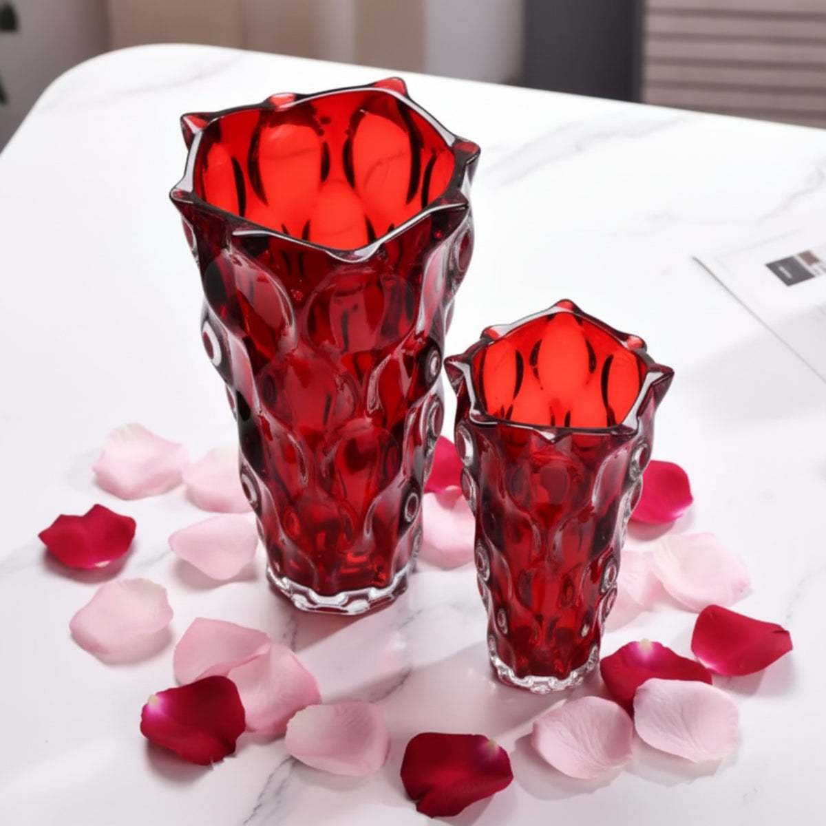 Two red textured glass flower vases of different sizes on a white marble tabletop scattered with pink and red rose petals.