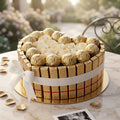A heart-shaped gourmet cake displayed on a marble table, decorated with white roses, gold-wrapped chocolates, and a gold chocolate bar wrapped around the side with a white ribbon bow. The background is a softly blurred outdoor garden setting.