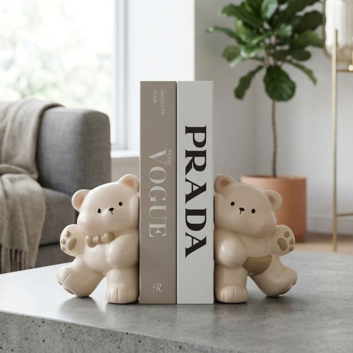 Two cream-colored bear-shaped bookends holding up two books on a gray tabletop in a modern living room setting. One book is beige with 'VOGUE' printed on it, and the other is white with 'PRADA' printed on it.