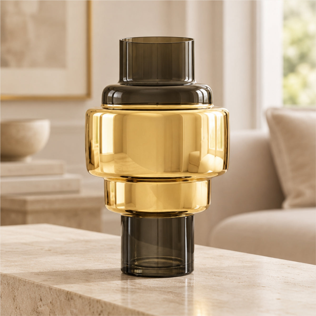A modern stacked glass vase with a reflective gold center section and dark smoky gray top and bottom sections, displayed on a light stone table in a softly blurred living room setting.