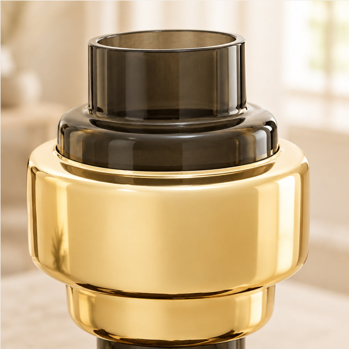 A close-up of a modern, tiered glass vase or candle holder with a shiny gold metallic lower section and a dark smoky gray glass upper section, set against a softly blurred warm indoor background.
