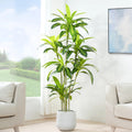 A tall artificial green plant with long, narrow leaves in a light gray cylindrical pot, placed in a bright living room between two beige armchairs.