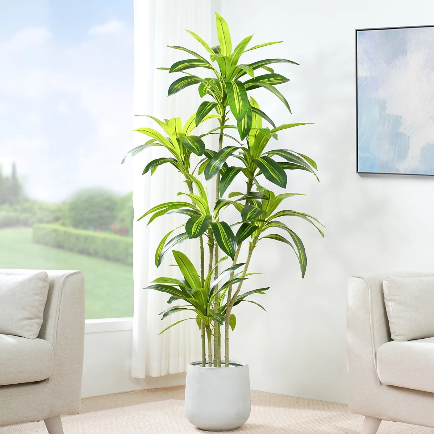 A tall artificial green plant with long, narrow leaves in a light gray cylindrical pot, placed in a bright living room between two beige armchairs.