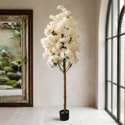 An artificial cherry blossom tree with dense white flowers and green leaves, standing in a black pot on a light wood floor in a bright room with a mirror and window.