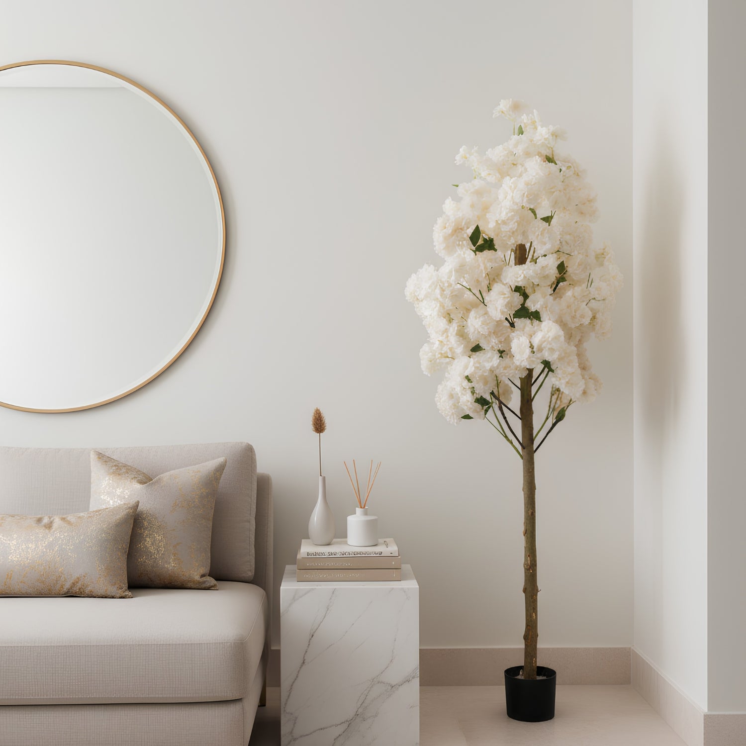 A tall artificial flowering tree with dense white blossoms and a brown trunk in a black pot, placed beside a light beige sofa and a white marble side table in a softly styled room.