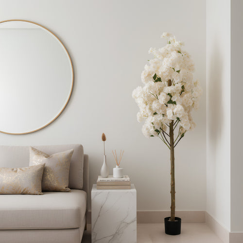 A tall artificial flowering tree with dense white blossoms and a brown trunk in a black pot, placed beside a light beige sofa and a white marble side table in a softly styled room.