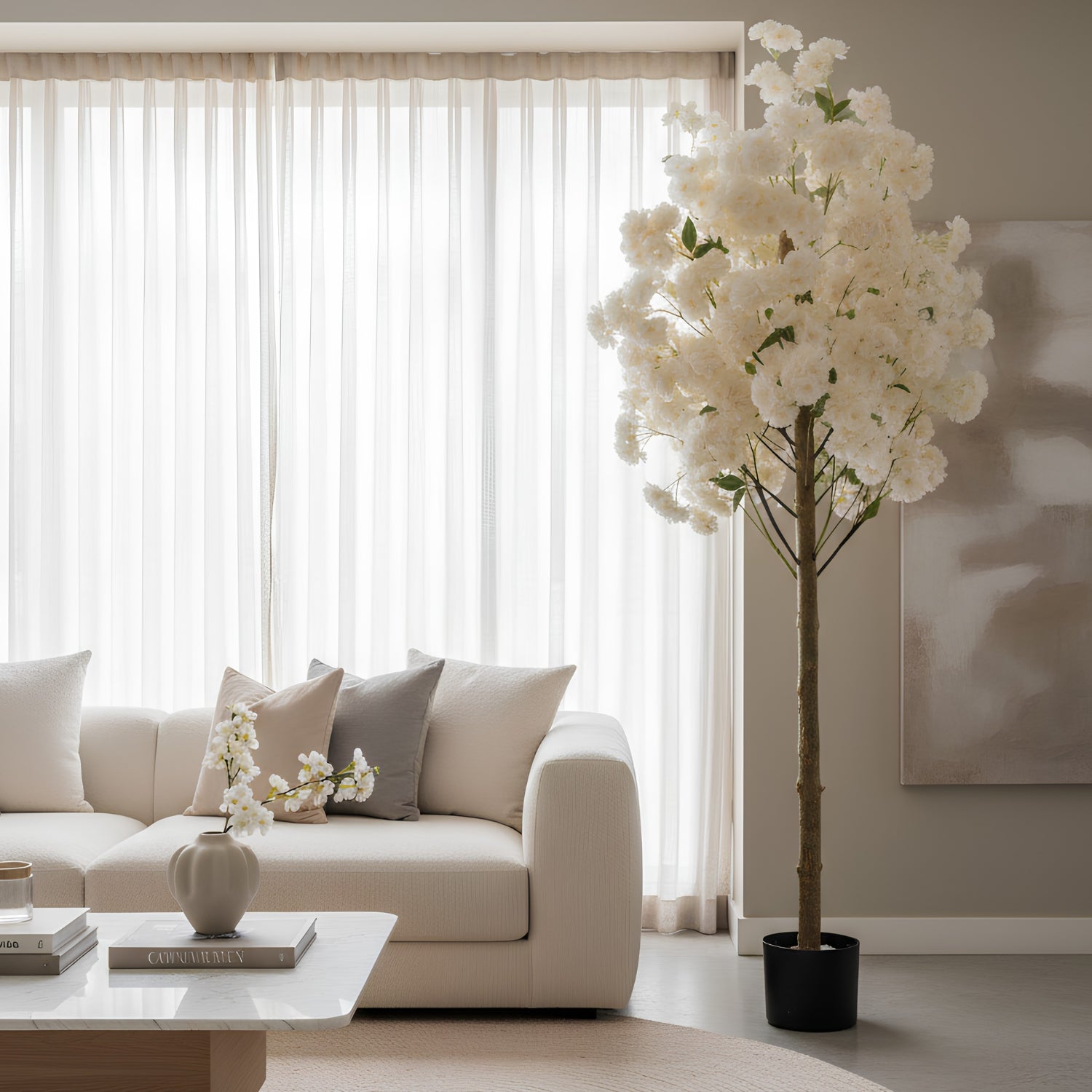 A tall artificial flowering tree with dense white blossoms and a brown trunk stands in a modern living room beside a cream sofa and sheer white curtains.